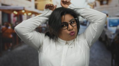 Gözlüklü bir kadın şehir terasında, canlı bir şehir ortamında neşeli bir ifade sergileyen tavşan kulaklı bir jest yapıyor..