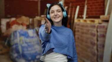 Tuğla ve malzemelerle çevrili bir inşaat sahasında kulaklık takan bir kadın elini uzatarak dinamik ve neşeli bir sahne yaratıyor..