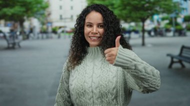 Sıcak kazaklı genç İspanyol kadın yeşil ağaçlar ve banklarla dolu kalabalık bir caddede dikilirken, pozitiflik ve açık şehir manzarası izlenimi veriyor..