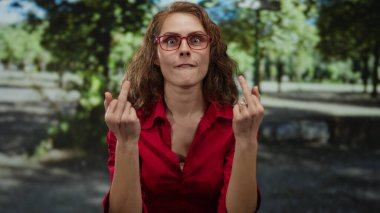 Gözlüklü bir kadın, güneşli bir park ortamında meydan okuyor. Kırmızı bir tişört giyiyor. Yeşil ağaçlarla çevrili jestler yapıyor.