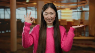 Otelde pembe kazaklı bir kadın gülümseyerek el hareketi yapıyor, rahat Asya atmosferinde zarif Çin tarzını yakalıyor..