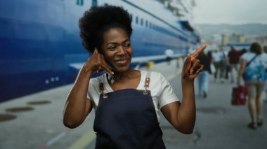 Limandaki kadın önlük giyerken el kol hareketi yapıyor, arka planda deniz olan büyük bir teknenin yanında duruyor, boş zamanların bir karışımını somutlaştırıyor ve açık deniz sahnesinde çalışıyor..