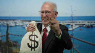 Takım elbiseli kıdemli bir adam elinde dolar işareti olan bir para çantası tutuyor. Arka planda botlarla deniz kenarında güvenle gülümsüyor ve yolsuzluğa işaret ediyor..