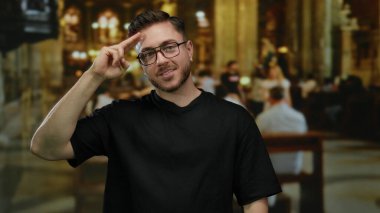 Kilisenin içinde sakallı genç adam jest yapıyor, gözlüklü ve siyah gömlekli, loş ışıklandırma altında sıralarda oturan insanların yumuşak arka planlarında güvenle gülümsüyor..