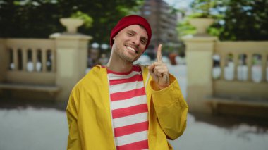 Sarı ceketli ve kırmızı bereli genç adam, şehir sokaklarında gülümsüyor, neşeli ve neşeli bir hava sergiliyor, bulanık şehir manzarası var..