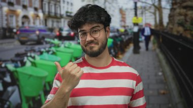 Çizgili tişörtlü, sakallı genç İspanyol adam yeşil scooterlı ve bulanık şehir geçmişli bir şehir caddesinde başparmak kaldırıyor..