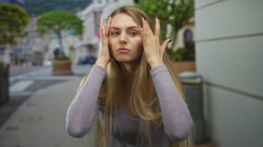 Uzun saçlı bir kadın aydınlık bir günde yeşil ve mimariyle çevrili günlük bir bluz giyerken saçını düzeltiyor..