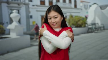 Kırmızı giyinmiş, açık havada gülümseyen, kendini sıcak tutan, güneşli bir sokakta mutluluk ve memnuniyet gösteren bir kadın..