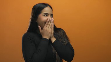 Elleri ağzının üzerinde gülen bir kadın canlı turuncu bir duvarın karşısında neşeli, neşeli bir atmosferi olan, neşeli ve izole bir ortamda neşe ve şaşkınlığı ifade ediyor..