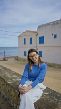 Canlı mavi bir kazakla gülümseyen bir kadın taş bir duvara yaslanmış, Mallorca 'nın kıyı kasabasında güneşli bir günün tadını çıkarıyor, arkasında beyaz binalar ve mavi panjurlar var..