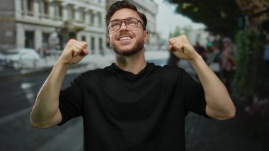 Sakallı ve gözlüklü genç adam bir şehir caddesinde başarıyı kutluyor, kendine güveni tam bir ifade sergiliyor..