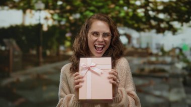 Şehir caddesinde elinde kurdeleli pembe hediye kutusu olan bir kadın, mutlu bir şekilde gülümsüyor gözlük ve süveter takıyor, bulanık şehir geçmişiyle çevrili..