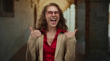 Tarihi bir üniversite koridorunda el kol hareketi yapan, şaşırdığını ve merakını dile getiren, kırmızı gözlük takan ve kapalı bir akademik ortamda bej ceket giyen bir kadın..