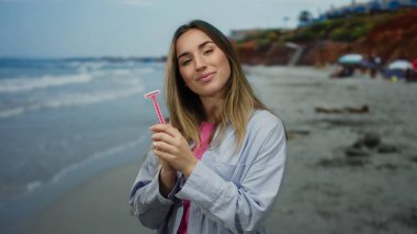 Güneşli bir günde elinde jiletle gülümseyen bir kadın canlı bir deniz arka planına sahip neşeli açık hava aktiviteleri sergiliyor..