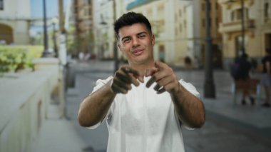 Genç adam güpegündüz hareketli bir şehir caddesinde rahat bir şekilde gülümsüyor günlük beyaz bir gömlek giyiyor ve kentsel bir ortamda olumlu ve ulaşılabilir bir atmosfer yaratıyor.