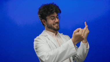 Sakallı, beyaz önlüklü genç adam orta parmaklarıyla mavi bir arka plana doğru esprili hareketler yapıyor. Gülümsüyor ve neşeli ama isyankar bir tavır sergiliyor..