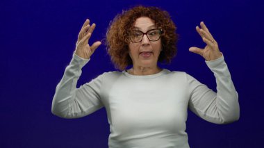 Woman gesturing humorously with expressive face against blue background wearing glasses and white shirt conveying fun emotion in isolated setting