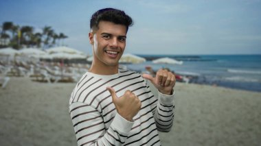 Genç adam kumsalda gülümsüyor, çizgili gömlek giyiyor, arka planda deniz ve şemsiyeler var, baş parmaklarını kaldırıyor, rahat bir sahil havası yakalıyor..
