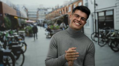 Canlı bir şehir caddesinde el çırpan genç bir adam gözlüklü ve arka planda insan ve bisikletli bir süveter giyen canlı bir şehir çevresi sergiliyor..
