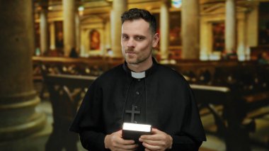 Kilise ortamında, elinde İncil, inanç ve dini bağlılık simgesi olan siyah cübbe ve çapraz kolye ile güvenle gülümseyen bir rahip..