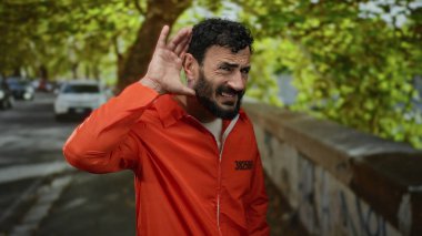 Turuncu tulumlu, sakallı bir adam caddede dikilip yeşil yaprakların ve arabaların arasında dikkatle dinliyor..