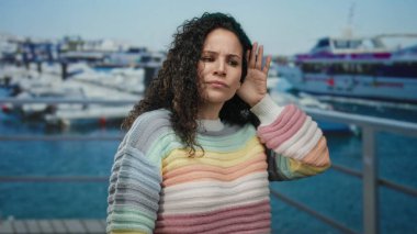 Renkli kazaklı genç İspanyol kadın güneşli bir günde arka planda teknelerle deniz kenarında jest yapıyor..