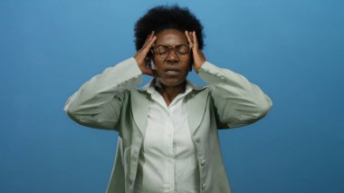 Kafası kapalı bir kadın, izole edilmiş mavi arka planda stresi tasvir ediyor, gözlük takıyor ve resmi ceket giyiyor afro saç stili ve güçlü duygular ifade ediyor..