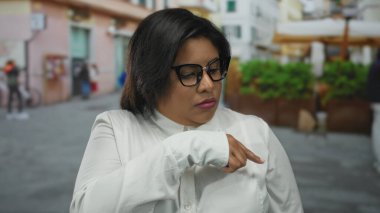 Gözlüklü, iri yarı Latin bir kadın canlı bir şehir caddesinde düşünceli bir şekilde el kol hareketi yapıyor, açık havada çeşitlilik ve güven gösteriyor..