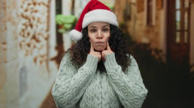 Noel Baba şapkalı ve kıvırcık saçlı genç bir kadın dışarıda şen şakrak bir sokakta, rahat bir kazakla, tatil ruhunu ve mevsimlik modasını sergiliyor..