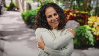 Renkli bir şehir caddesinde sıcak bir şekilde gülümseyen bir kadın, canlı çiçeklerle çevrelenmiş bir ortamda kendini rahatlatan bir sarılma veriyor..