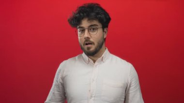 Sakallı ve gözlüklü İspanyol bir adam, canlı kırmızı bir arka plana karşı şaşkınlık ve neşe ifade ediyor, açık renk bir gömlek giyiyor, genç ve hayat dolu bir enerjiye bürünüyor..