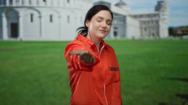 Turuncu hapishane tulumu giymiş bir kadın İtalya 'daki Pisa Kulesi' nin önünde el kamerasına uzanıyor. Hapishaneyi ikonik mimariyle harmanlayan eşsiz bir sahneyi yakalıyor..