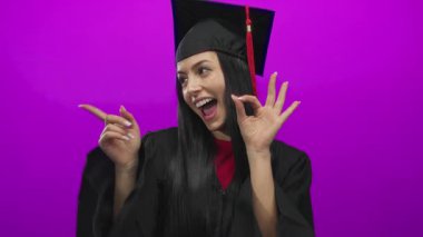 Mezuniyet şapkalı ve cüppeli genç İspanyol kadın pembe arka planda neşeli bir ifadeyle iyi bir jest yapıyor.