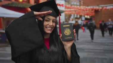 Mezuniyet cüppeli genç İspanyol kadın elinde Meksika pasaportu ve kalabalık bir caddede gülümsüyor..