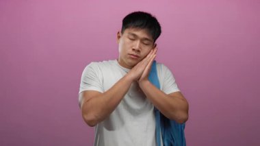 Sırt çantalı genç adam pembe arka planda uyuyormuş gibi yapıyor. Yorgunluk ve öğrenci yaşam tarzını somutlaştırıyor..