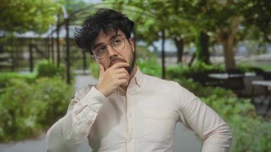 Sakallı ve gözlüklü genç adam ağaçlarla çevrili canlı yeşil bir parkta düşünceli bir şekilde el ele tutuşuyor..