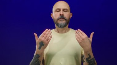 Man with beard and tattoos meditates against a blue wall showing focus and calm.
