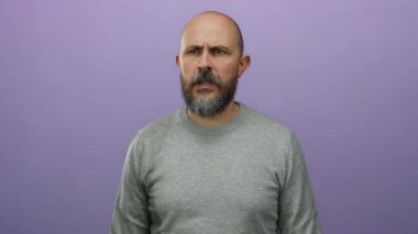 Gri kazaklı sakallı kel bir adam mor bir arkaplanı işaret ederek ciddi bir ifade sergiliyor..