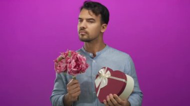 Genç bir adam elinde buket ve kalp şeklinde bir kutu tutarak canlı pembe arka planda, rahat bir gömlek giyiyor, romantik ve düşünceli tavırlar sergiliyor çiçekler ve hediye vurgusu yapıyor..