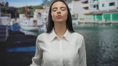 Beyaz tişörtlü bir kadın elleriyle kalp hareketi yapıyor ve canlı limanda öpüşüyor. Arka planda güneşli açık hava ortamının altında tekneler var..