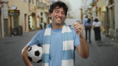 Sokakta elinde futbol topu ve kartlarıyla gülümseyen, mavi gömlek ve eşarp giyen neşeli bir şehir ortamının spor ve neşeli havasını somutlaştıran İspanyol bir adam..