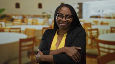 Kolları çapraz bir restoranda gülümseyen, gözlüklü ve sarı gömlekli bir kadın, arka planda ahşap sandalyeli ve masalı davetkar bir ortam sergiliyor..