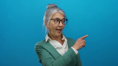 Gri saçlı, gözlüklü, gülümseyen yaşlı bir kadın mavi arka plana başparmak işareti veriyor. Pozitif olma ve onay öneriyor..