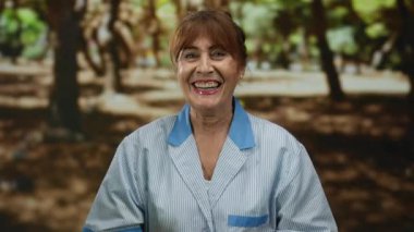 Parkta gülümseyen kıdemli kadın temiz bir üniforma giyerek açık hava arka planına karşı gerçek mutluluğu ve profesyonelliği gösteriyor..