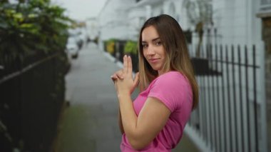 Şehir sokağındaki bir silah gibi parmaklarını şaklabanca gösteren, pembe bir gömlek giyen, açık şehir ortamında kendine güven ve yaratıcılık ifade eden bir kadın..