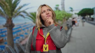 Genç sarışın kadın, güneşli bir sahil sahilinde gönüllü. Düşünceli ve düşünceli. Palmiye ağaçlarına ve mavi denize karşı kırmızı bir yelek giyiyor..