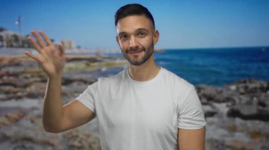 Genç adam kumsalda okyanusla ve güneşli gökyüzünün altındaki kayalarla el sallıyor, dost canlısı ve rahat bir hava iletiyor..