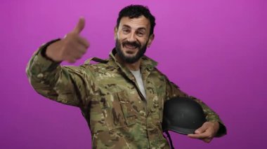 Askeri üniformalı İspanyol bir adam canlı pembe bir arka plana karşı miğfer tutarken olumlu ve hazır olduğunu bildiriyor..