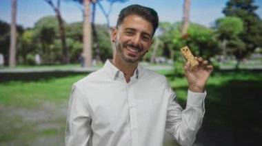 Beyaz tişörtlü genç İspanyol adam elinde granola çubuğuyla güneşli bir günde yeşil parkta gülümsüyor..