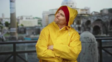 Sarı yağmurluk giyen ve kırmızı bereli bir adam gülümseyen ve eski Roma harabelerinde başparmak kaldıran şehir manzarası ve mimarisini sergileyen..